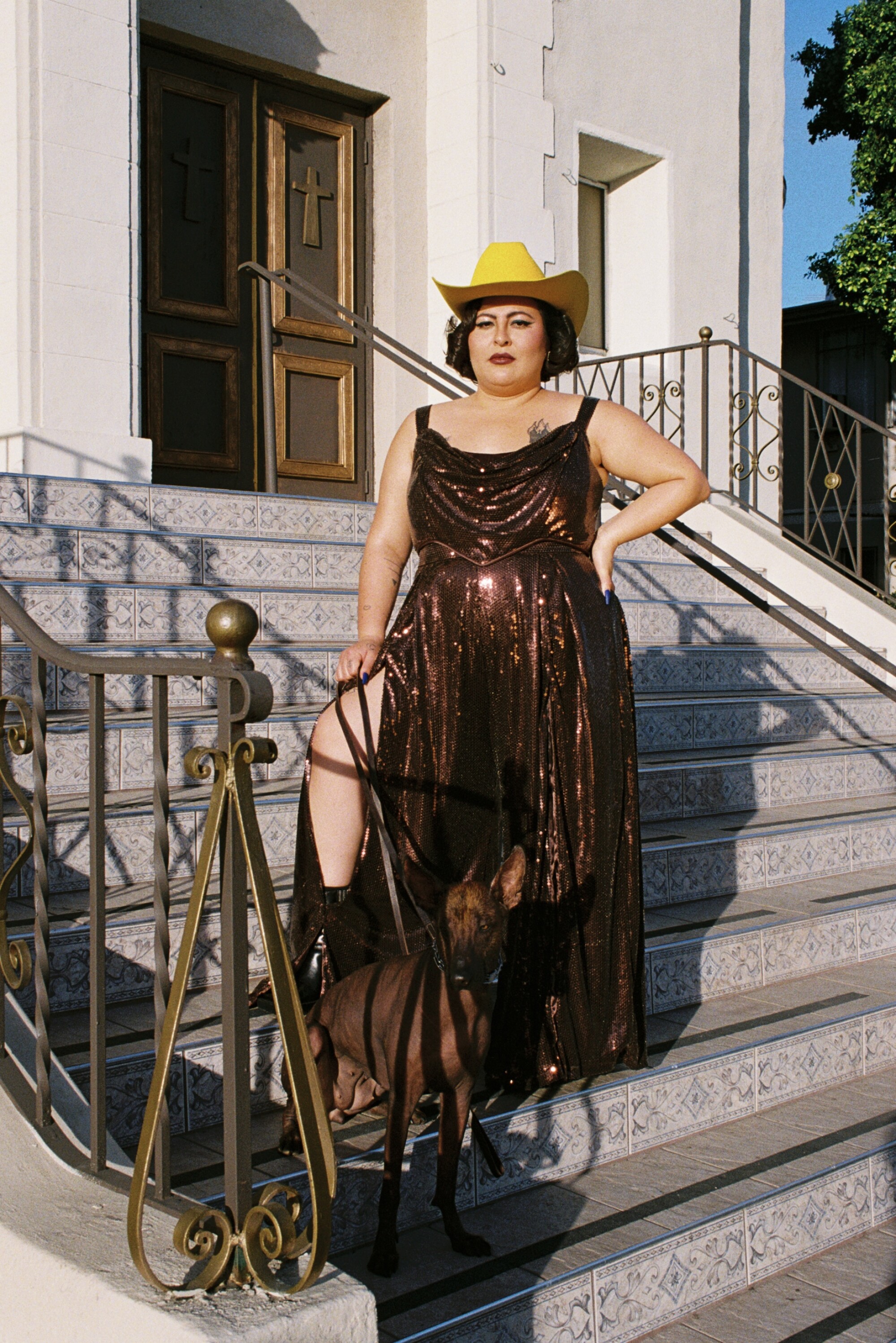 San Cha stands in front of a church with a dog in a brown sequined gown and yellow cowboy hat
