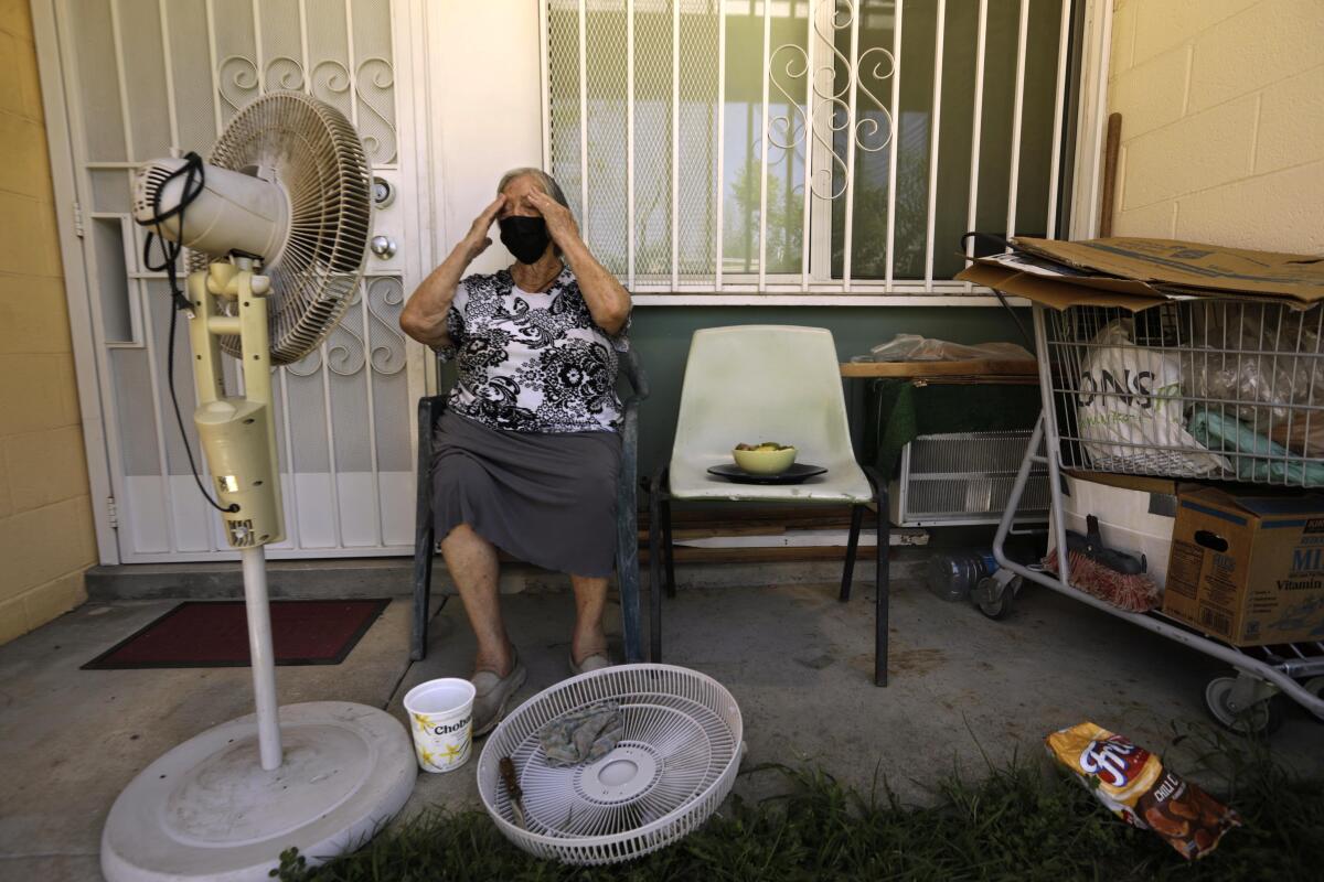 A woman wipes the sweat from her brow as she sits before a standing fan. 