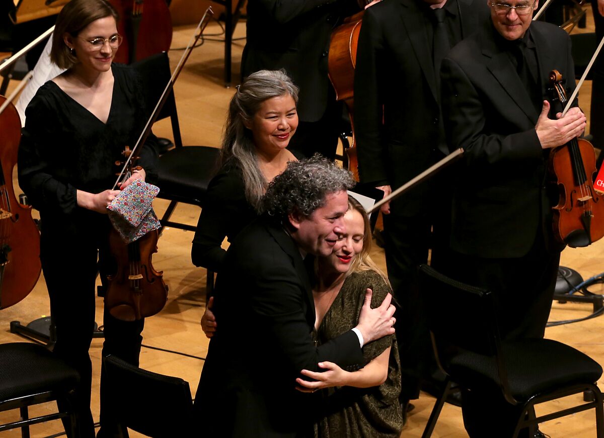 A conductor embraces a composer onstage as the orchestra's musicians look on and smile.