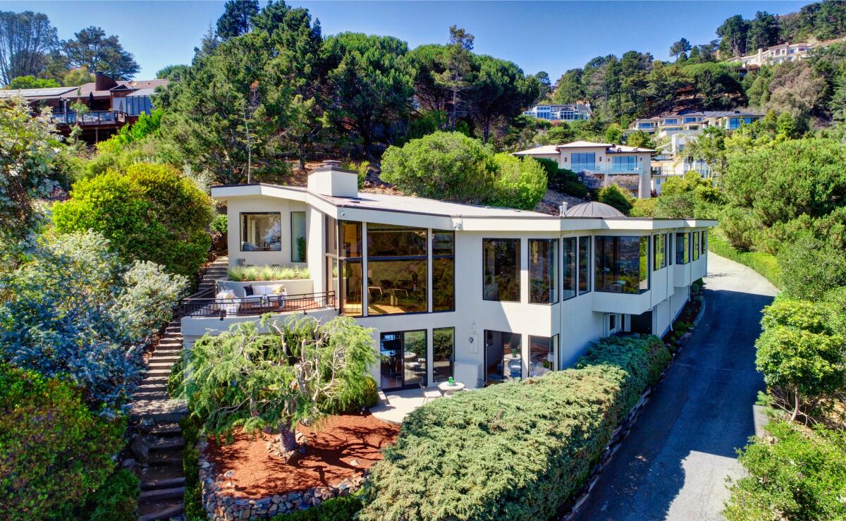 A two-story home with multiple large windows sits amid greenery on a hillside plot. 