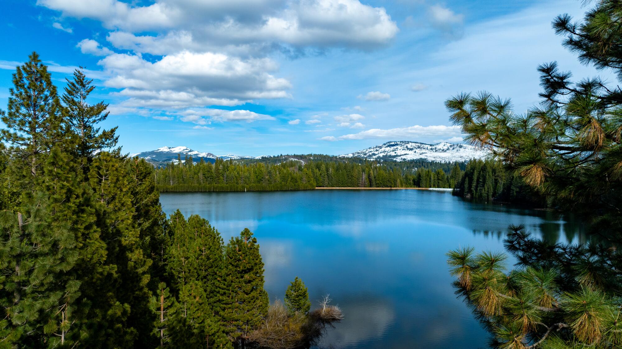 Clouds hover over a bright blue lake surrounded by evergreens. Snowy mountains rise in the background.