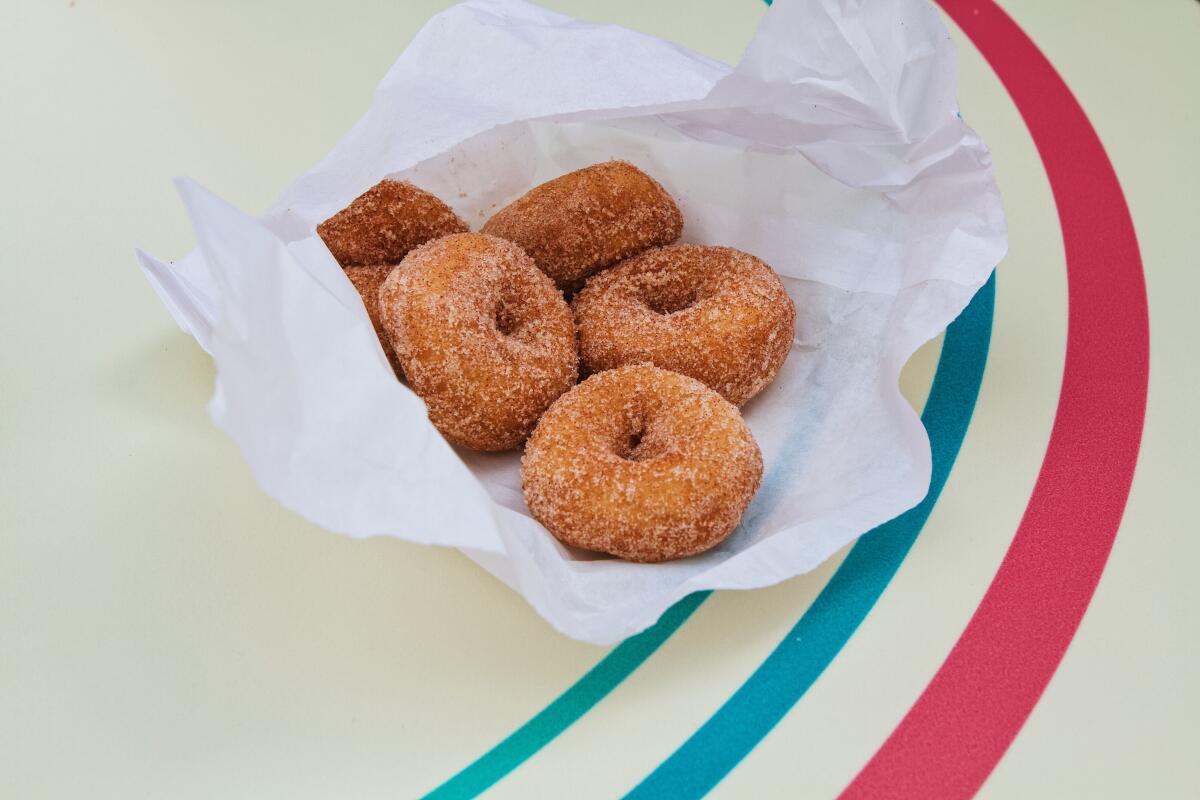 A white bag of six mini doughnuts dusted with cinnamon sugar sits on a table with blue and pink striping.