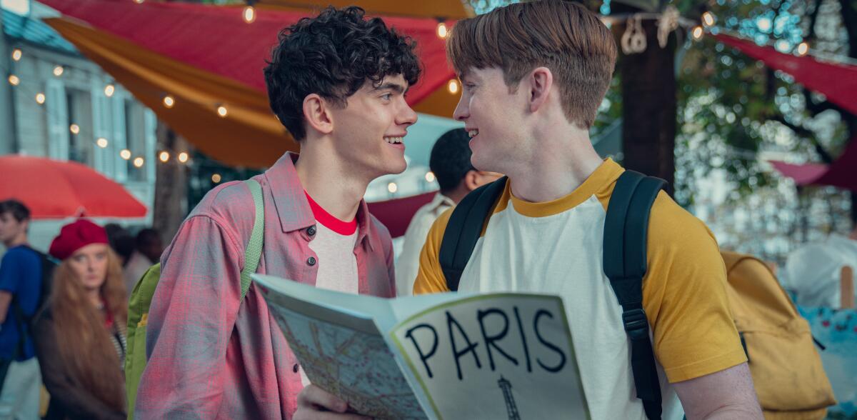Two young men stand outdoors look smiling at each other, one holding a pamphlet marked Paris