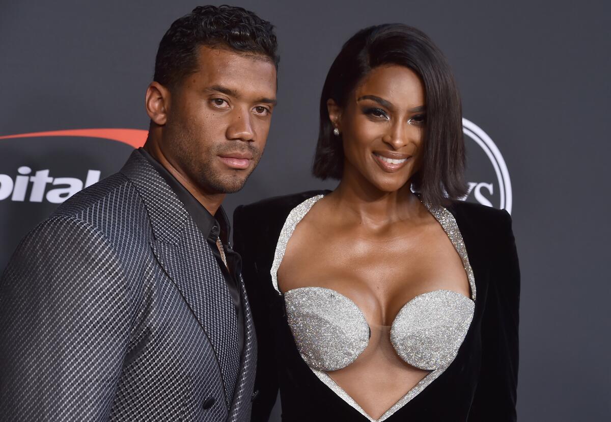 Russell Wilson, in a dark suit, stands next to Ciara, who wears a black dress with silver trim around a revealing cutout