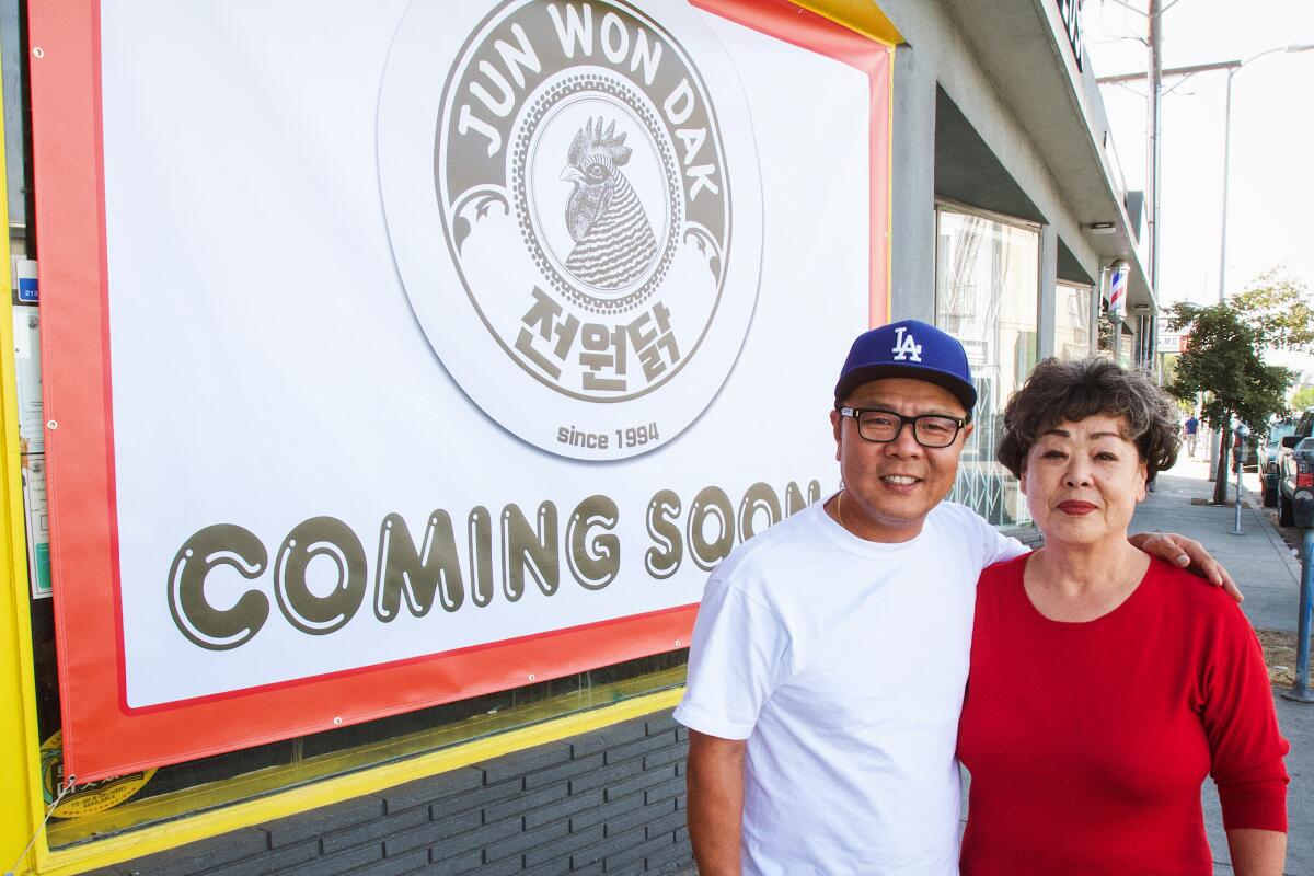 A man with his arm around a woman's shoulder, standing outside next to a large banner that reads "JUN WON DAK COMING SOON."