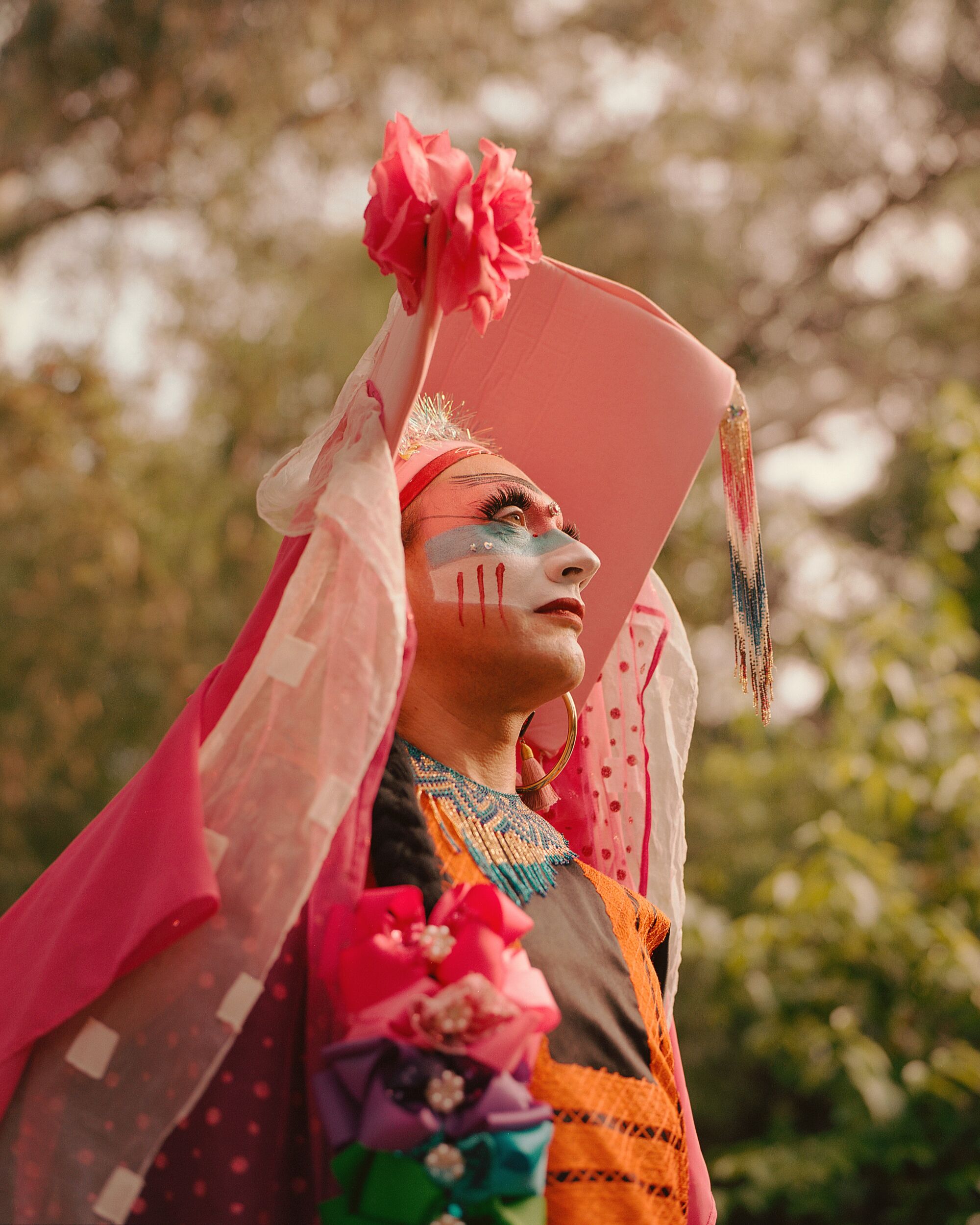 A drag nun in a peach colored dress with a colorful face standing outdoors looking up.