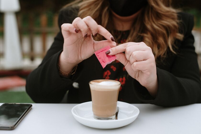 A young woman wearing a protective face mask adds a nonsugar sweetener to her coffee.
