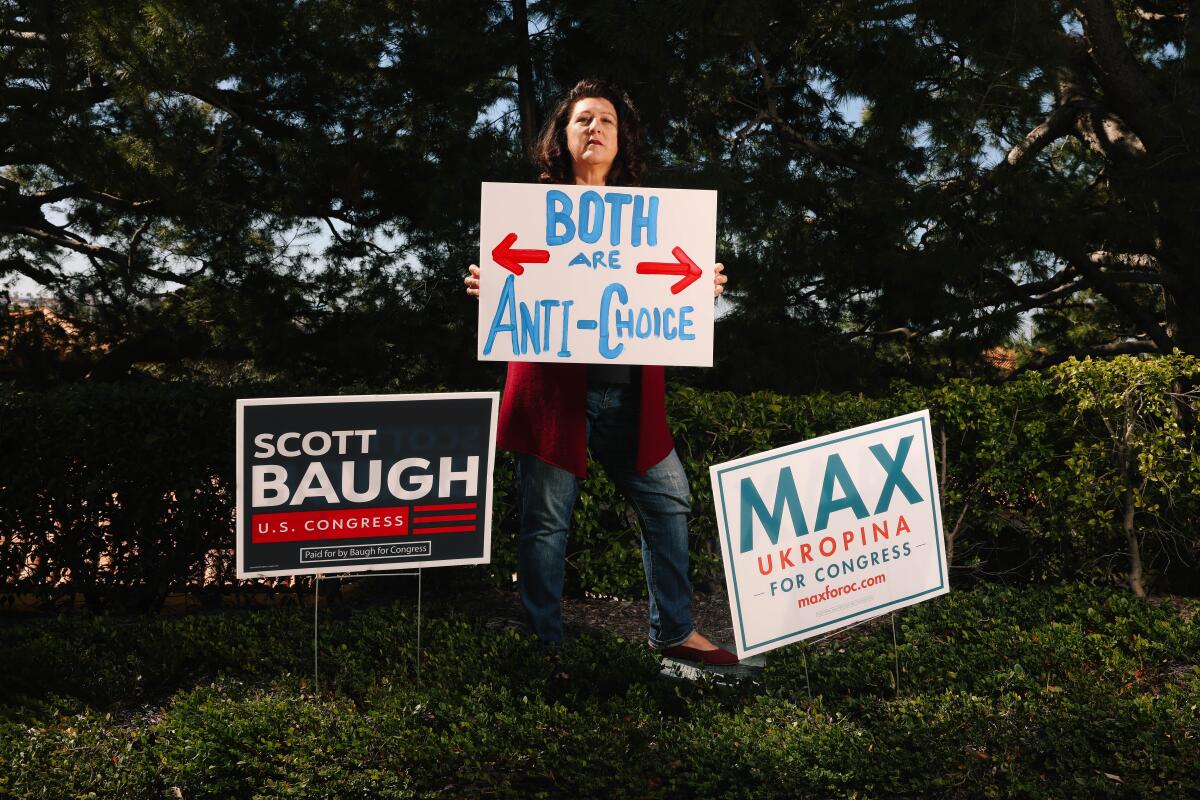 A woman poses for a portrait holding a pro-choice sign while standing between campaign signs for Republican candidates 