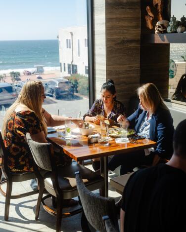 Three women sit at a restaurant table with an ocean view out the window next to them.