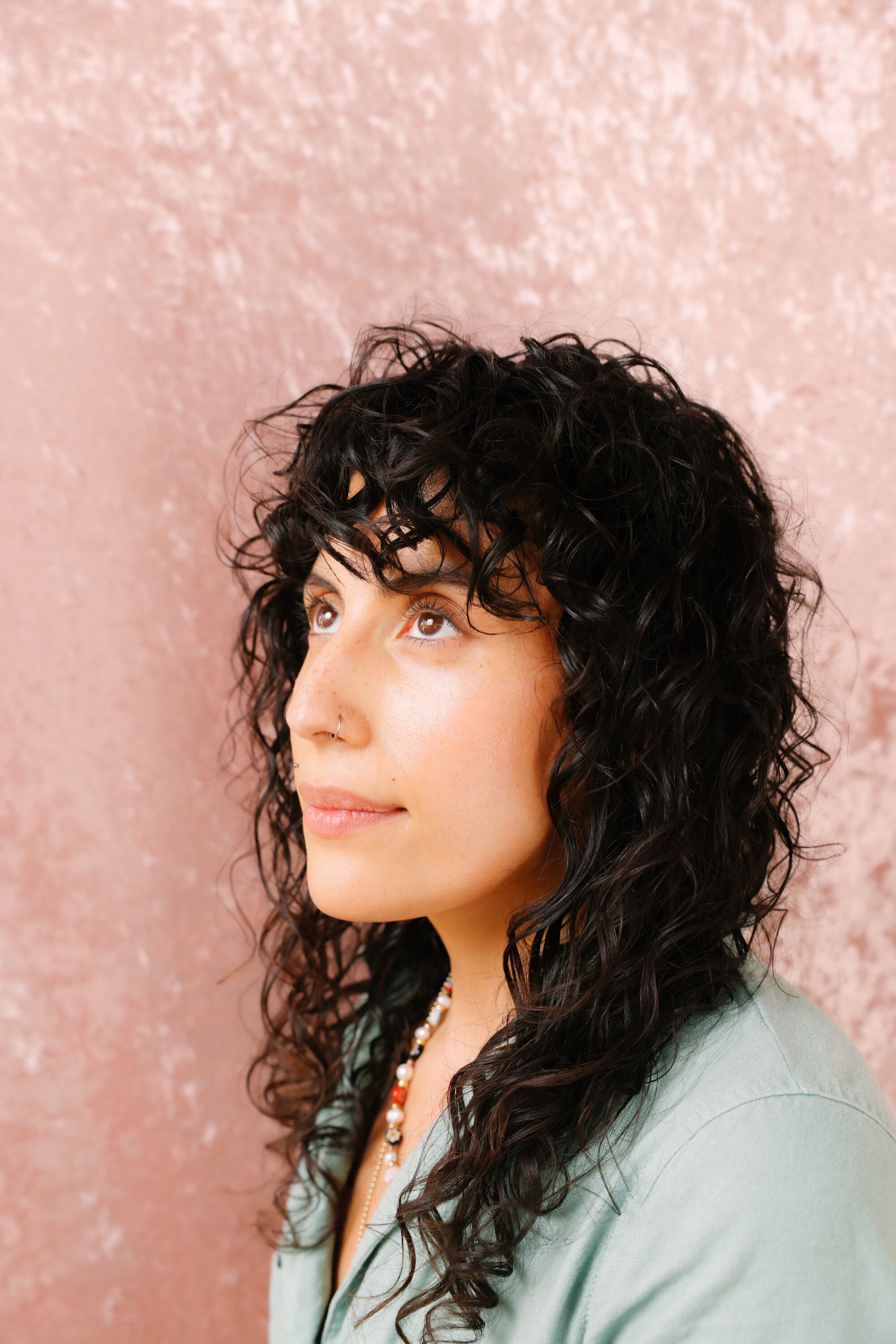 A person with curly long hair looks up for a portrait against a pink background.