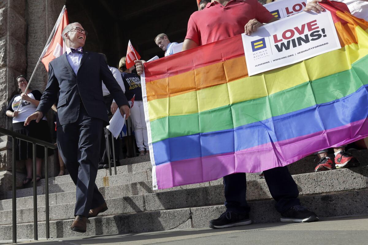 A man in a dark suit walks down a set of stairs near a person holding a rainbow banner that reads 