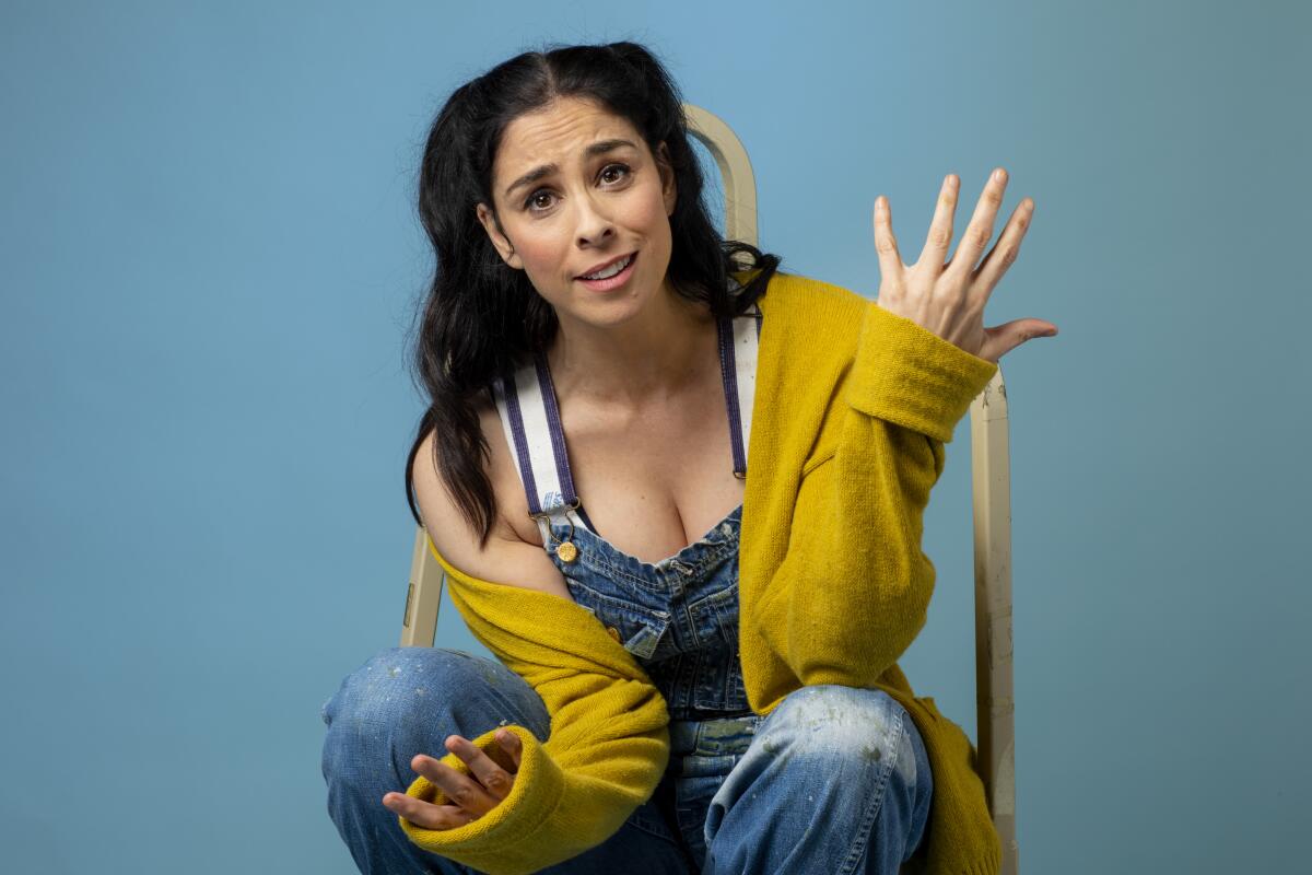 Comedian Sarah Silverman shrugs in a chair wearing overalls and a cardigan.