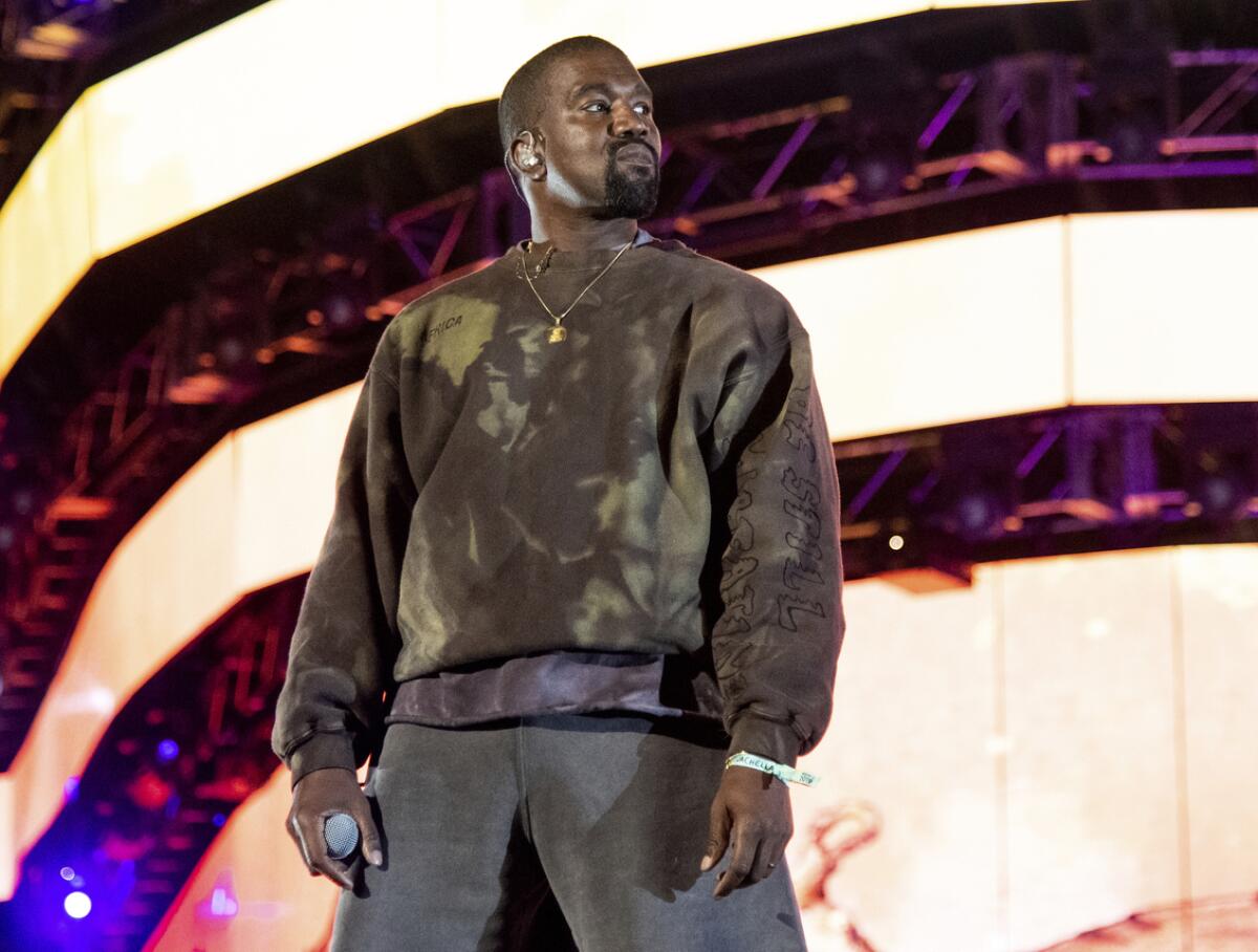 Kanye West wears a camouflage sweatshirt with gray pants as he holds a microphone while standing on a stage