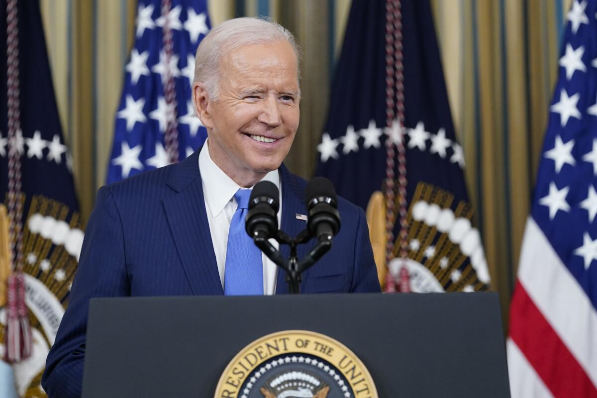 Smiling, President Biden stands at a podium with American flags in the background.