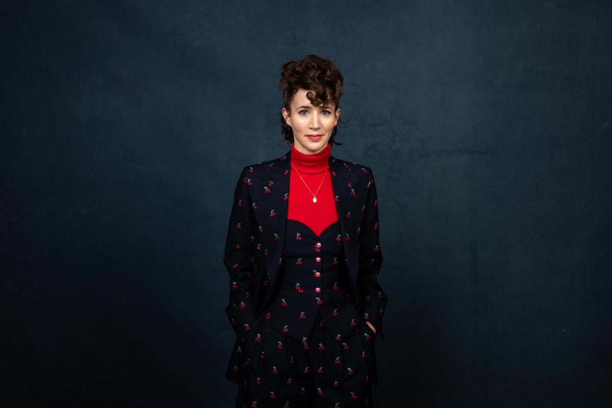 Miranda July, in a navy blue dress and matching blazer with cherries, stands in front of a blue backdrop.