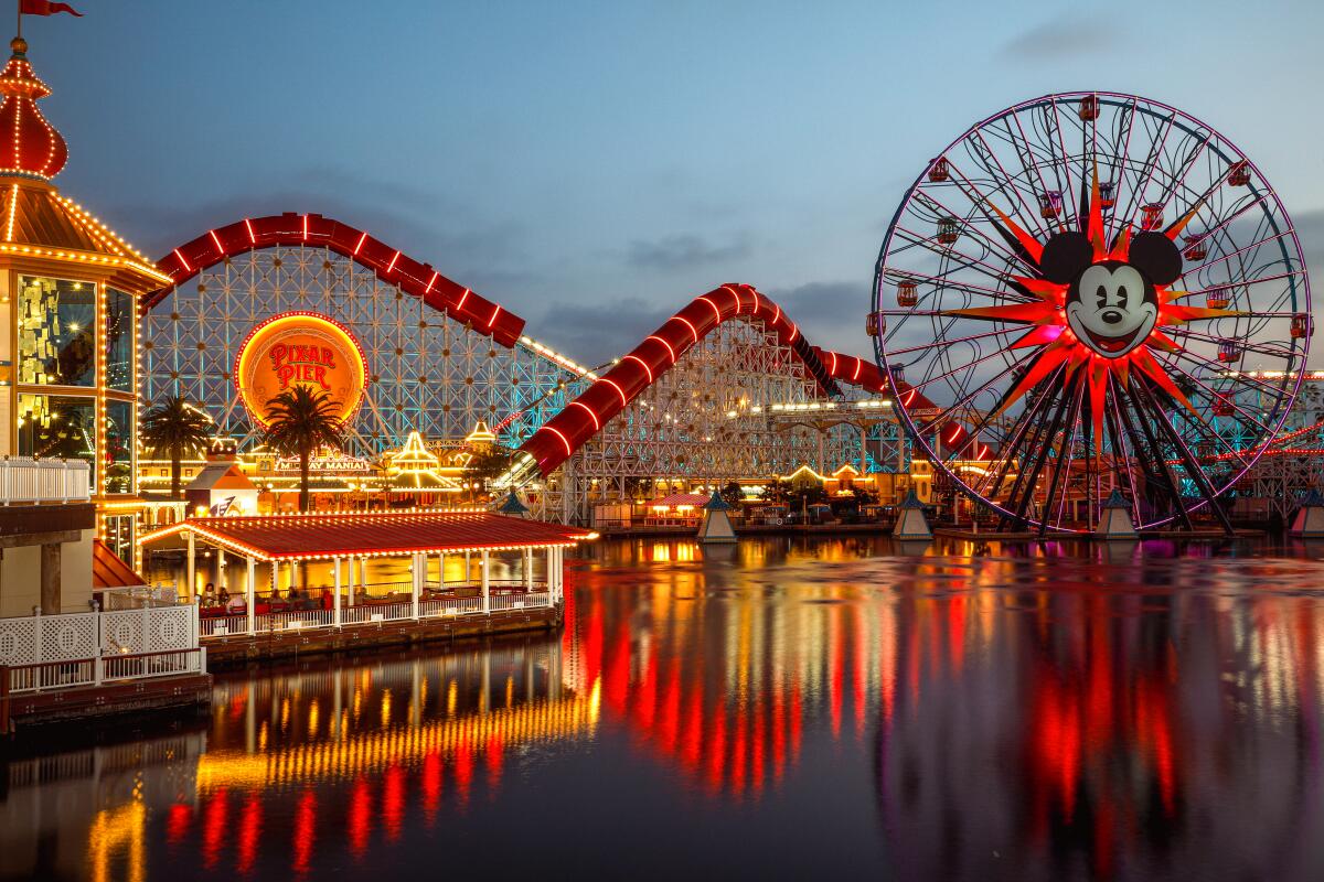 A smiling mouse's face is at the center of a Ferris wheel alongside a lighted-up coaster at Disneyland.