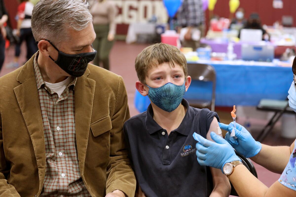 A father sits with his son as his son receives a COVID-19 vaccine from a nurse.