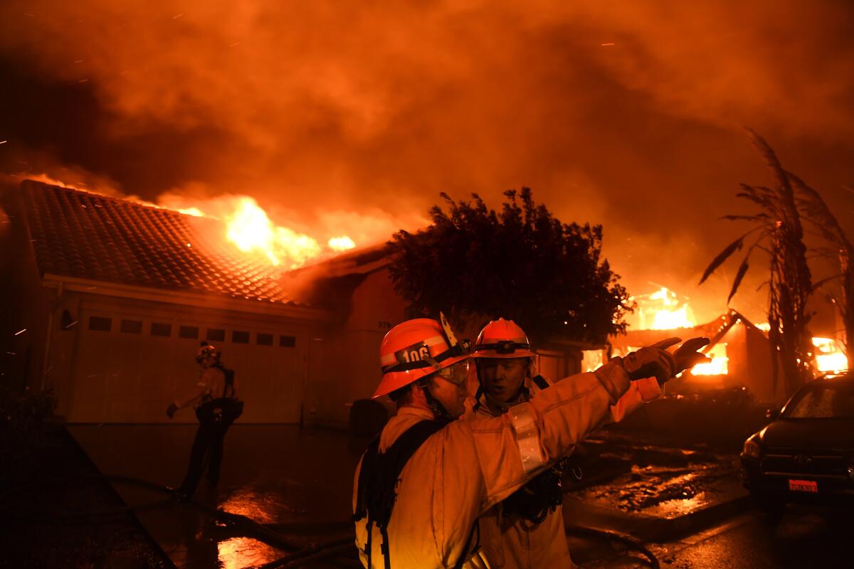 Two firefighters talk as flames consume houses behind them.