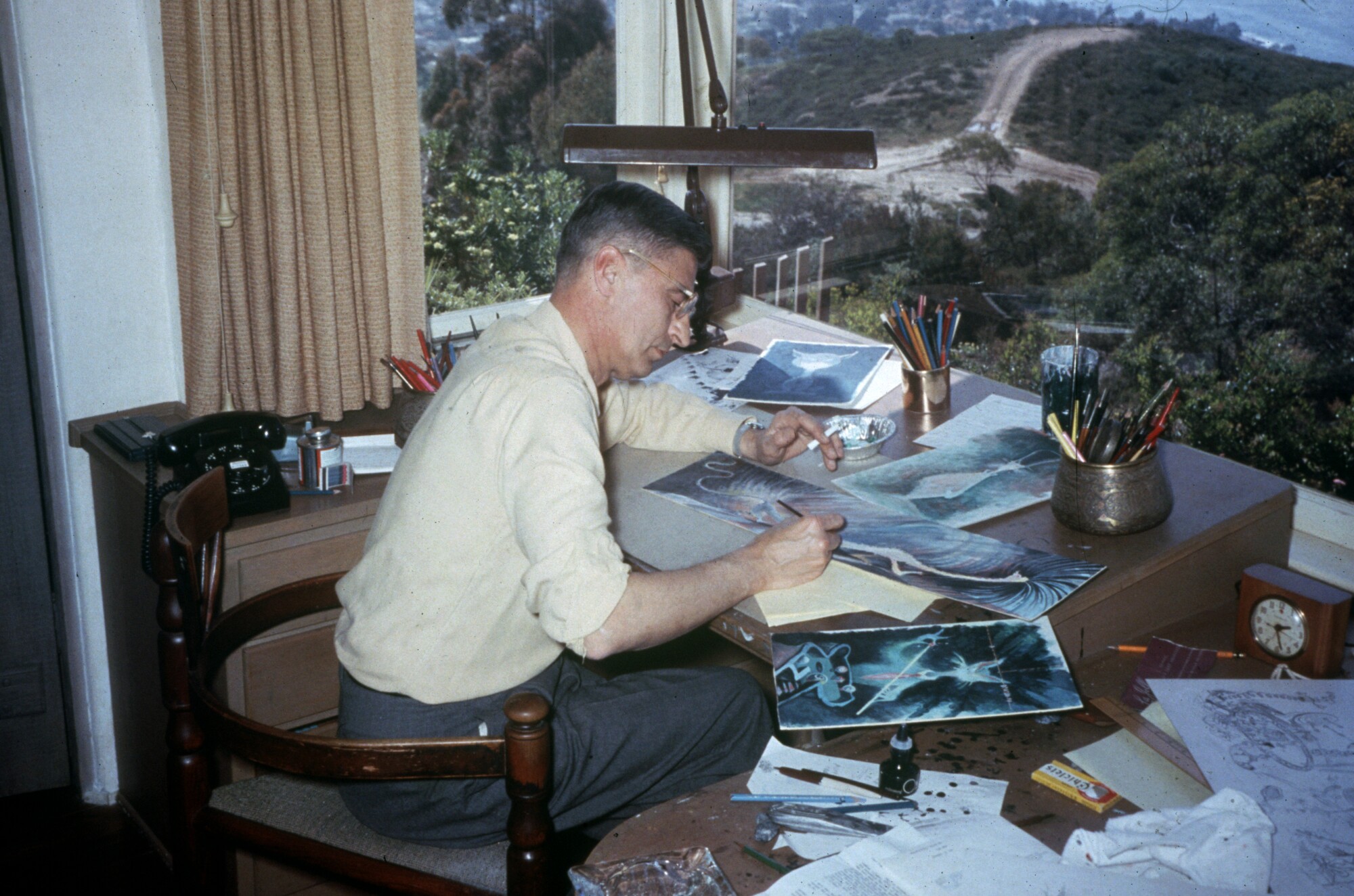 A man draws while sitting at a desk in front of a window that has a view of a slope and the ocean