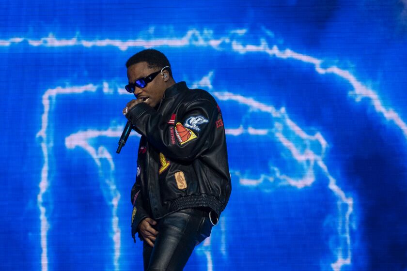 A man in black leather raps into a microphone in front of a brightly lighted background
