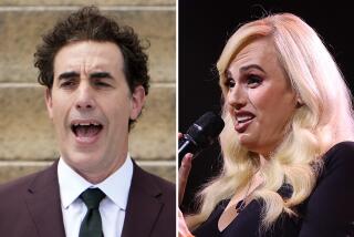 Sacha Baron Cohen in a brown suit and tie with his mouth open. Rebel Wilson in a black dress speaking into a microphone
