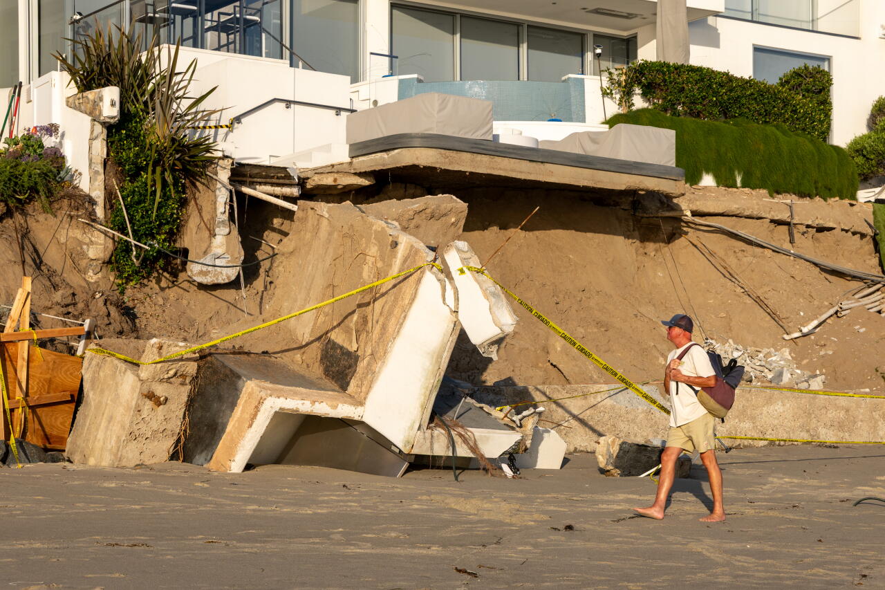 A man walks by damaged buildings on a beach.