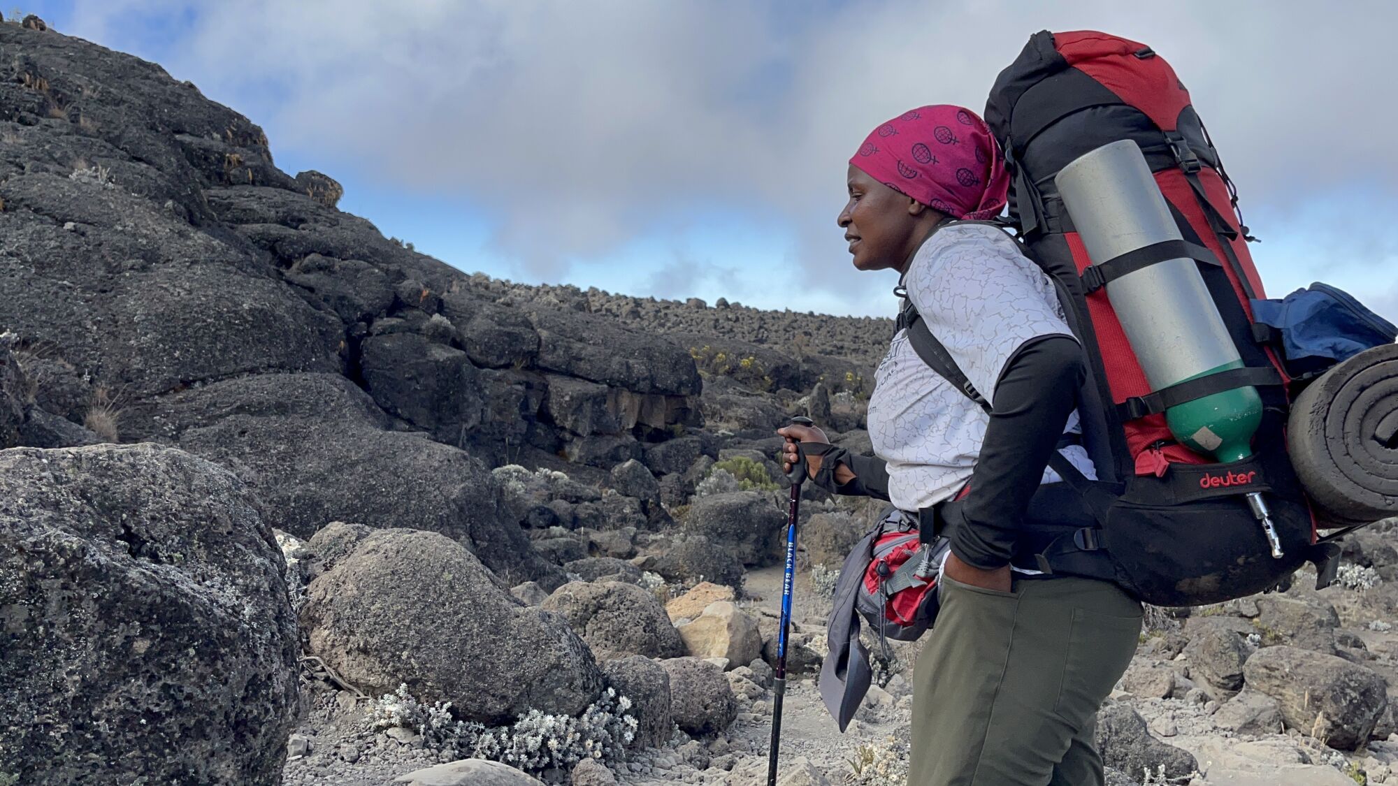 A woman in a red bandana holds a walking stick and carries a large red-and-black backpack in a mountain setting