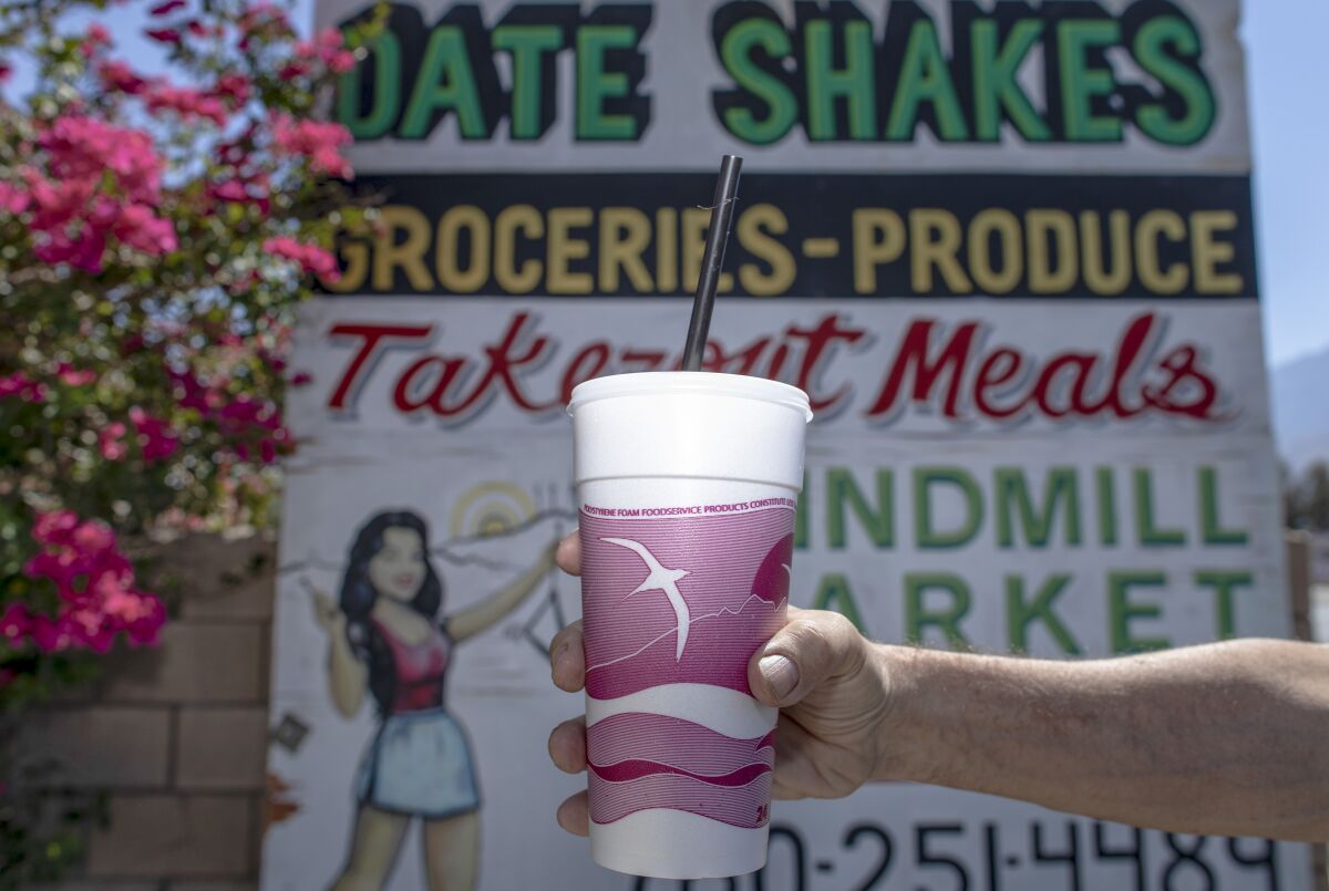 An outstretched arm holds a date shake in front of a sign advertising them.