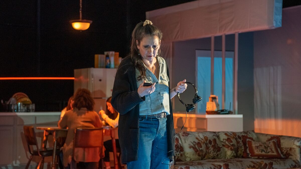 A woman stands holding headphones and looking at a cellphone, with people seated at a table behind her, in a play.