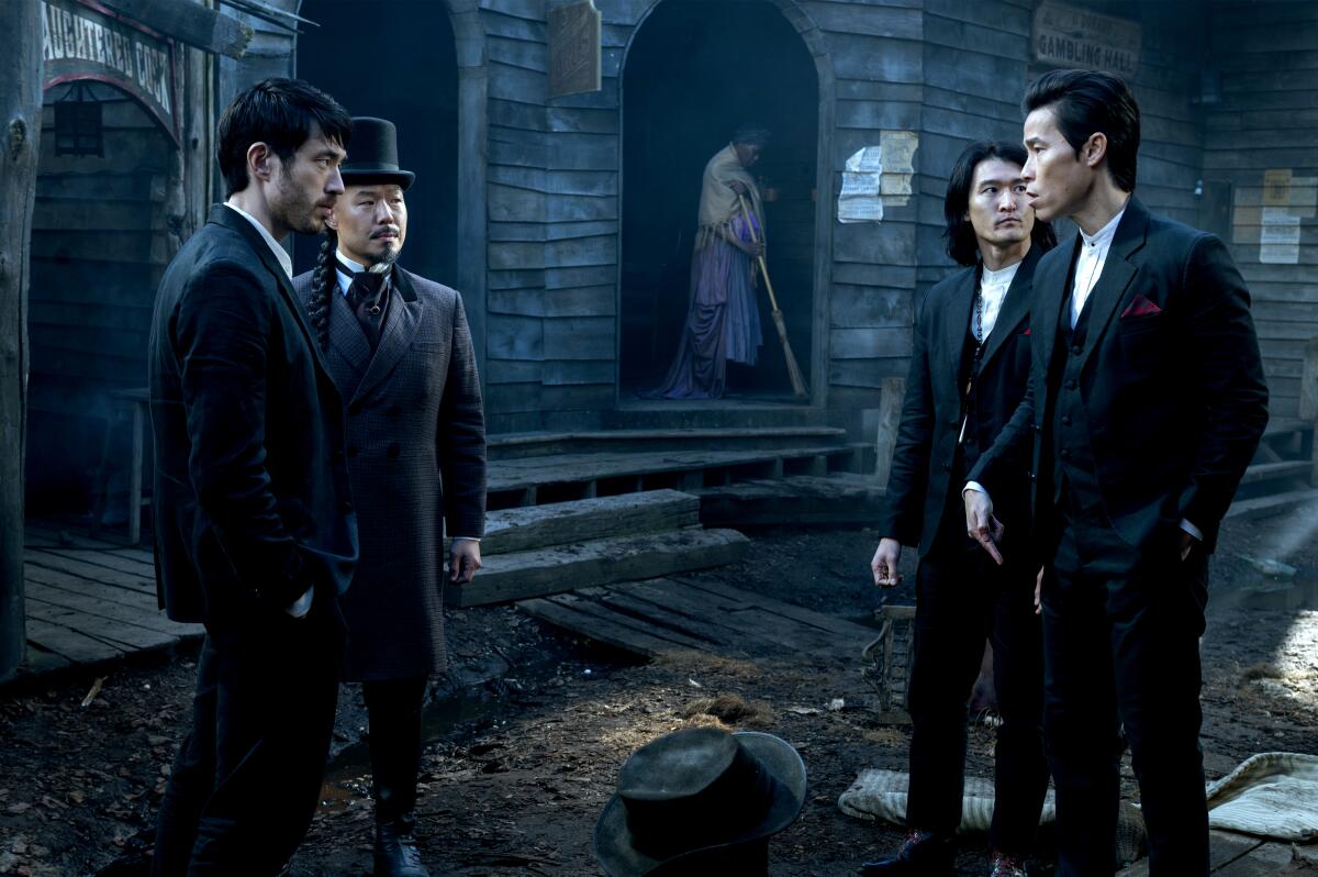Two men in dark suits stand next to each other, looking at two other men in suits, on a street with stone walls.