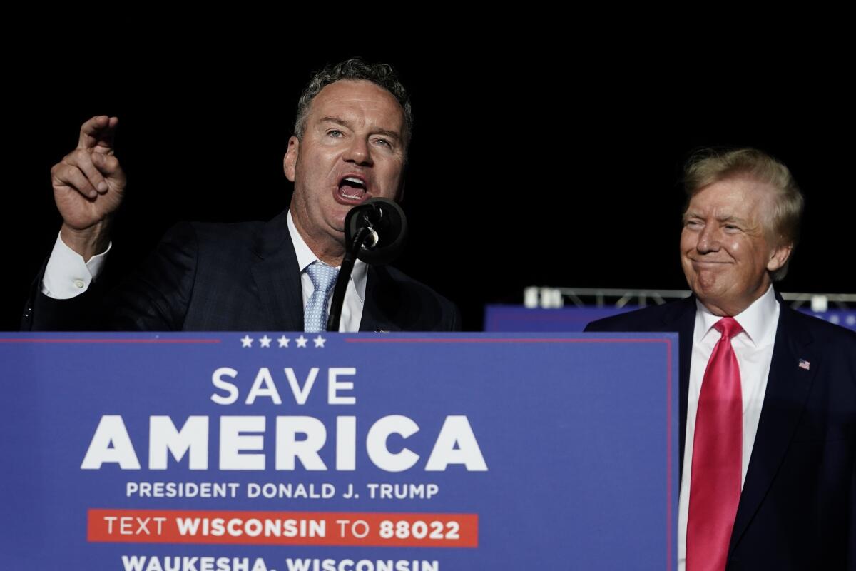Tim Michels speaks into a microphone behind a sign that says "Save America" as former President Trump, beside him, smiles. 