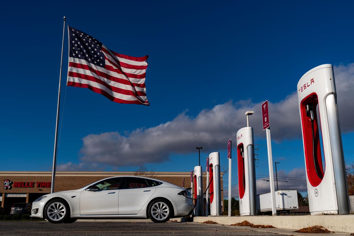 An electric vehicle charges at a Tesla Supercharger station as a U.S. flag flies above. 