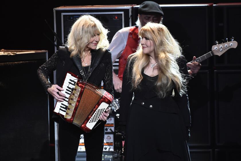 Two women with long blond hair in dark formal wear looking at each other while performing on a stage
