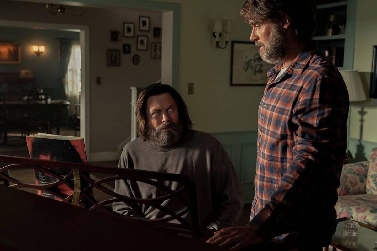 A man standing next to another man seated at a piano in "The Last of Us."