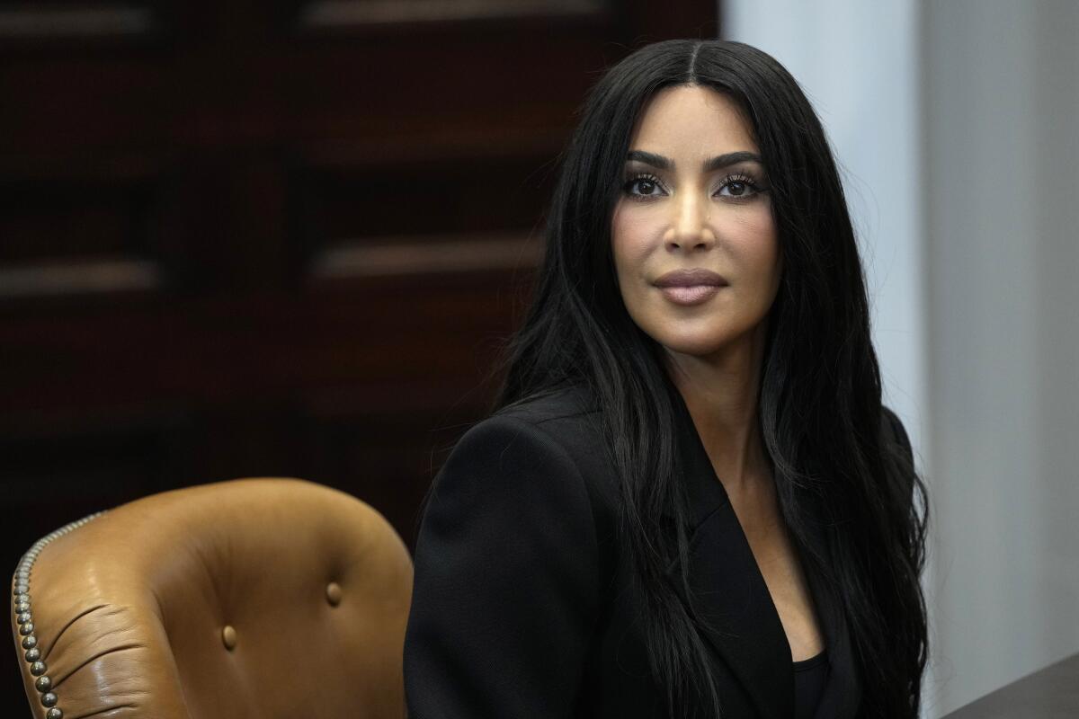 Kim Kardashian sitting alertly in a brown leather chair in a black jacket and top looking straight ahead