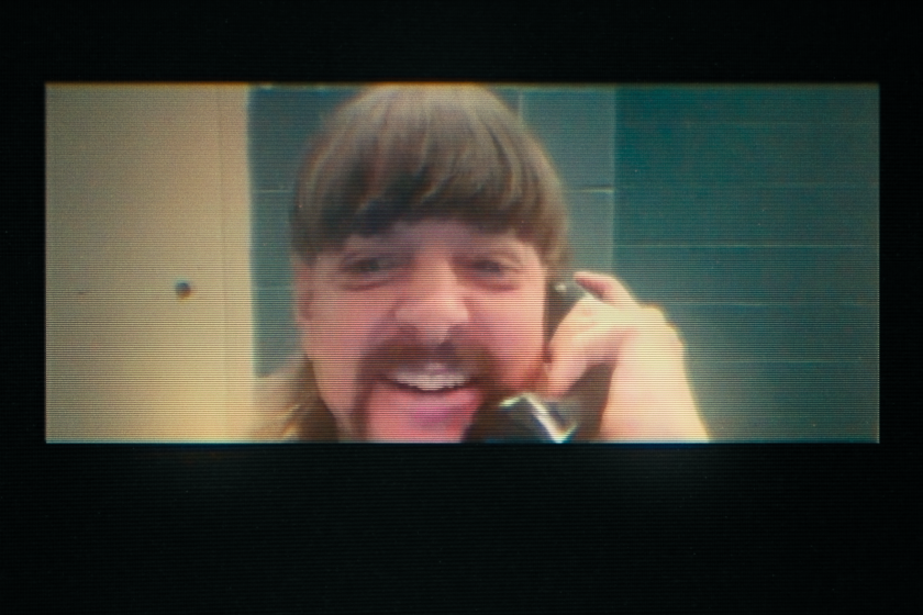 A grainy image of a man smiling while talking into a prison telephone