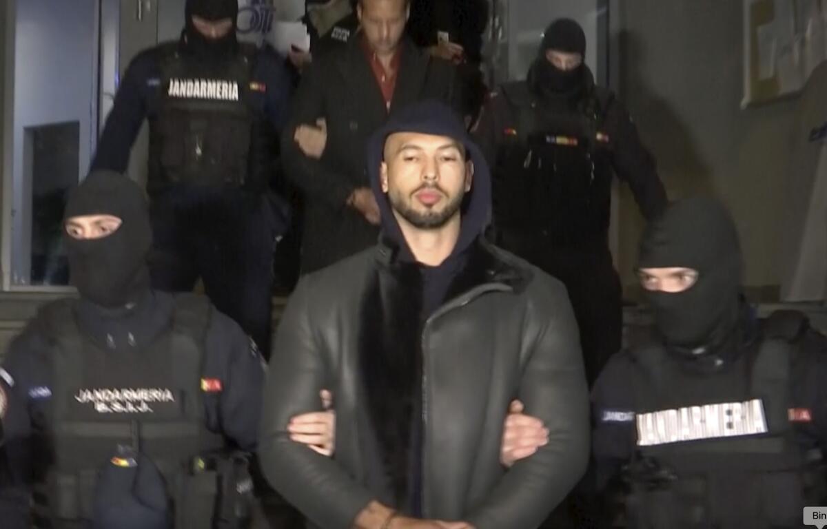 Man in black hoodie with trimmed black beard is led out by group of masked law enforcement wearing black vests