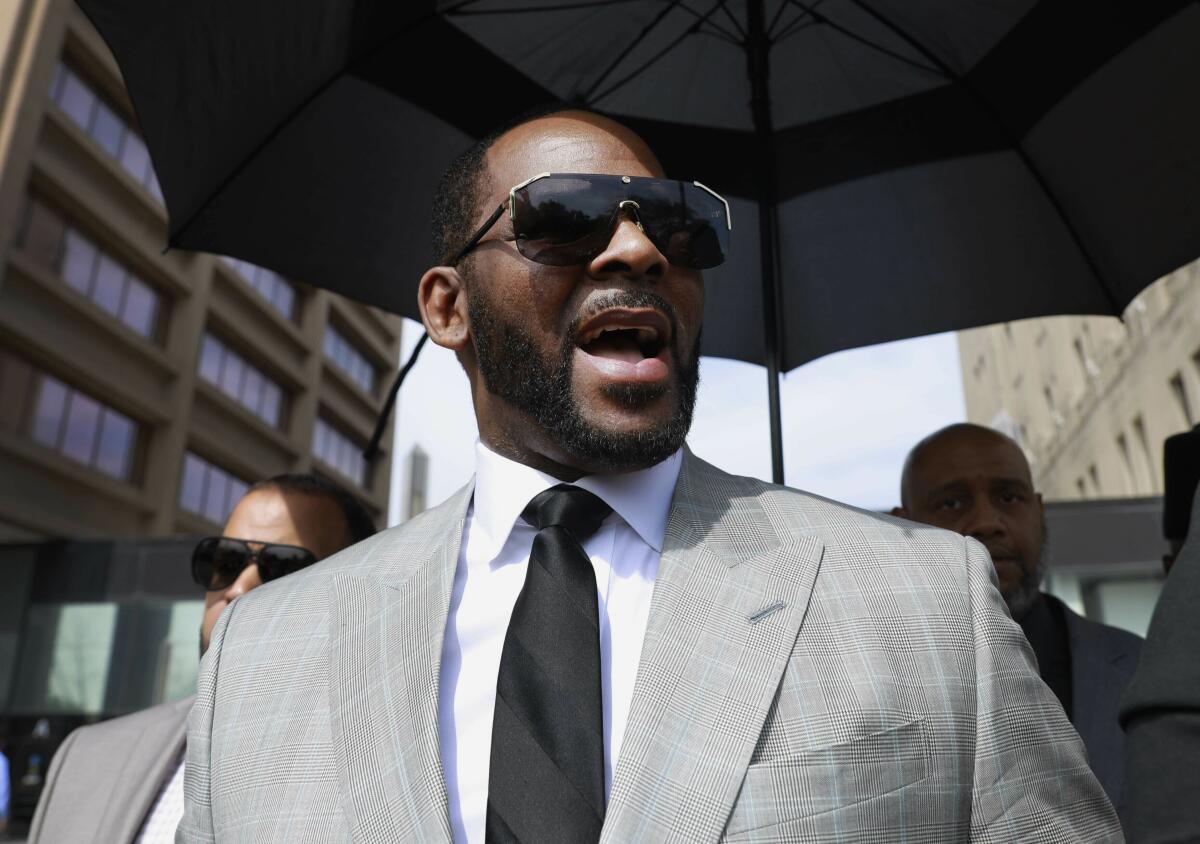 A man with his mouth open in a gray suit, black tie and sunglasses getting shaded by an umbrella