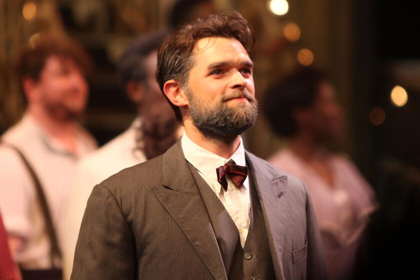 Chris Peluso smiling wearing brown suit and red bow tie while on stage, looking off at a crowd in front of cast