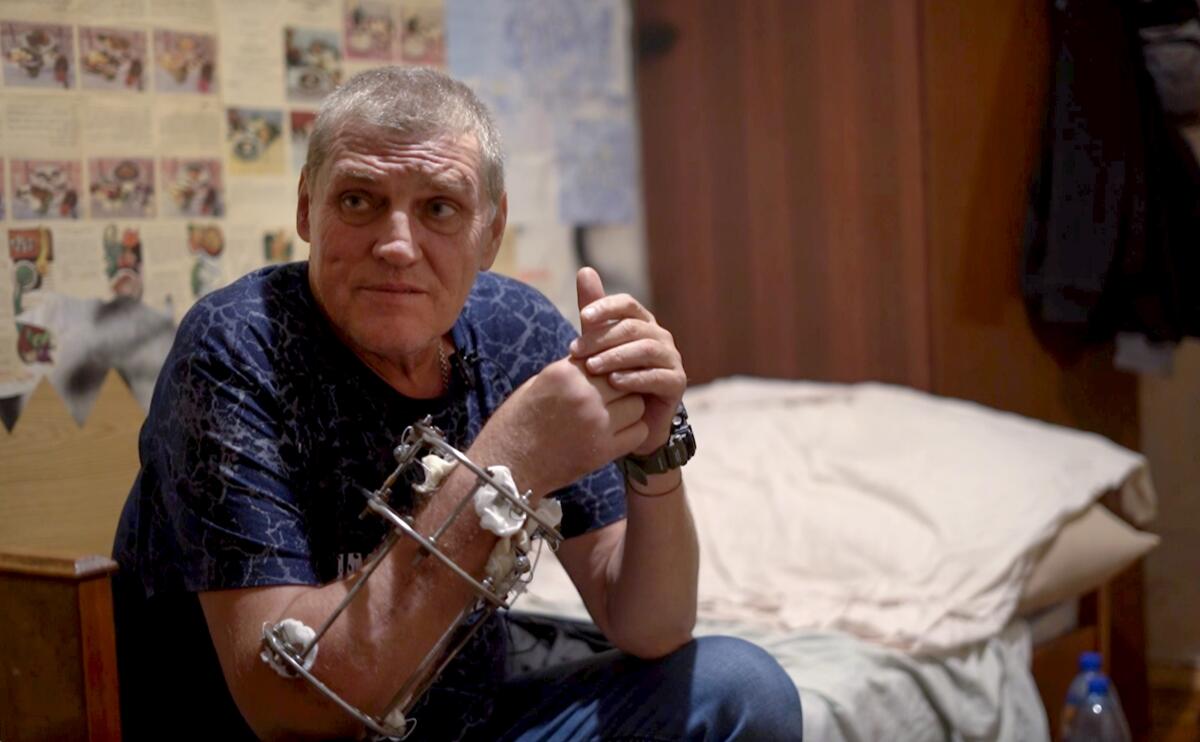 A man sits on the edge of a bed with a metal brace on his right arm