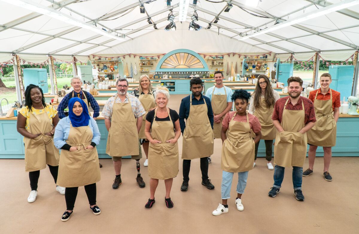 Twelve cast members of 'The Great British Baking Show,' all in tan aprons, stand in a large kitchen tent.