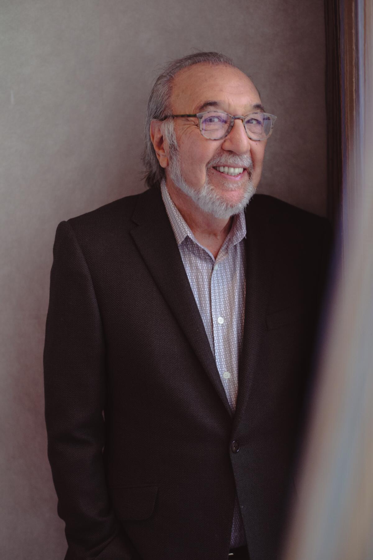 Man with a beard and glasses, dress shirt and jacket smiles and looks at camera with side-eye.