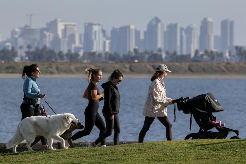 SAN DIEGO, CA - JANUARY 15, 2024: With the San Diego skyline in the background, a woman jogs past other women walking along a sidewalk at Crown Point Beach in San Diego on Monday, January 15, 2024.