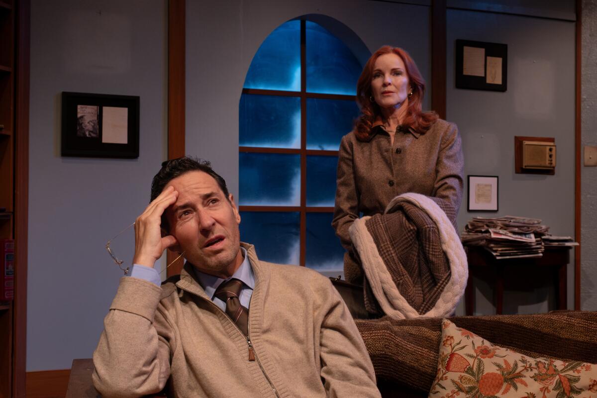 A worried-looking man sits on a couch, with a woman standing behind him holding a coat