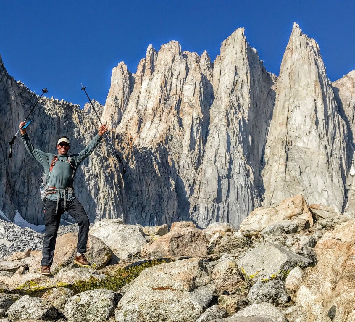 A man holding hiking poles raises his arms in a pose below a large rocky mountain peak.