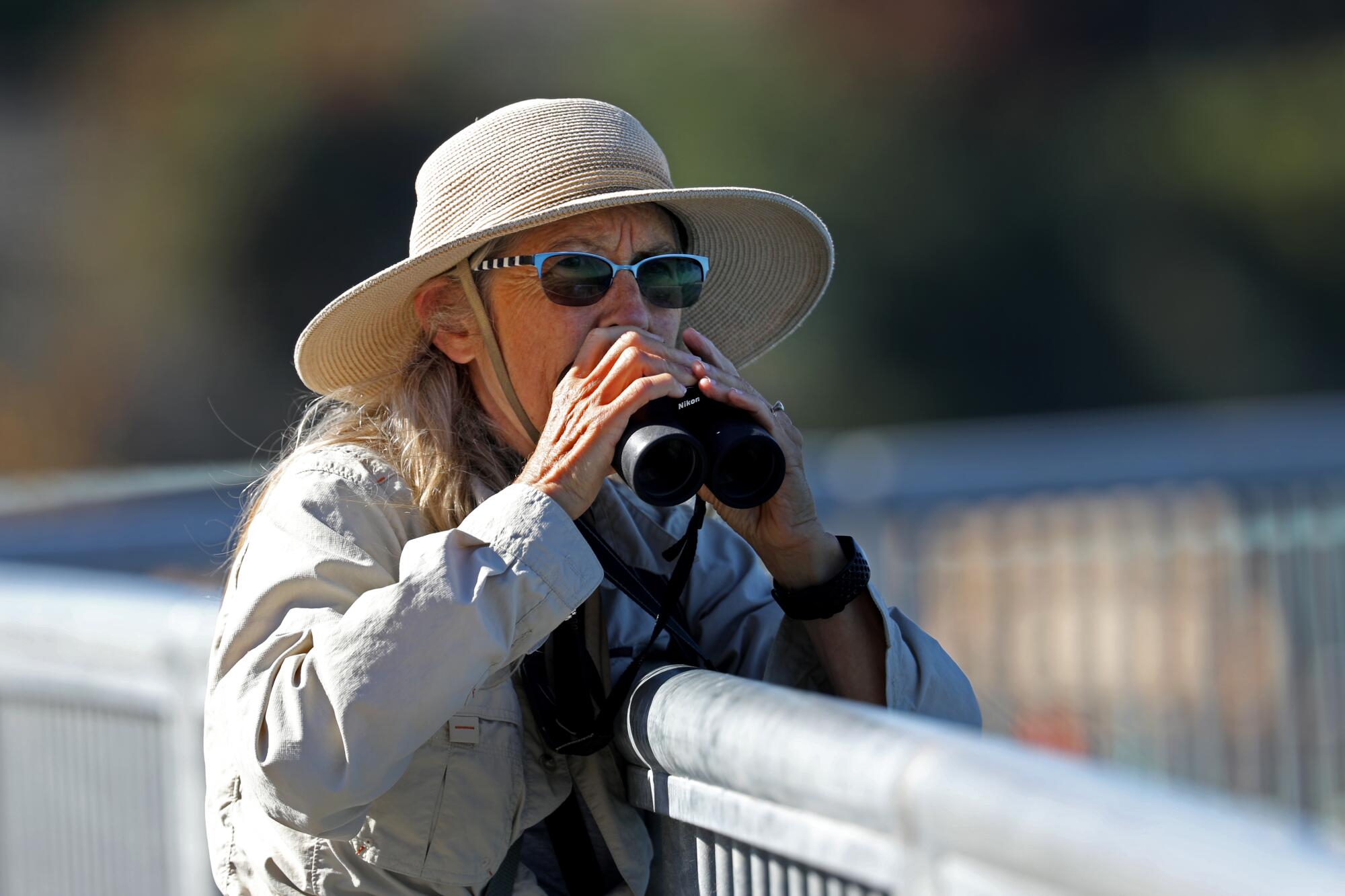 A woman in a sun hat and sunglasses holds binoculars near a metal fence 