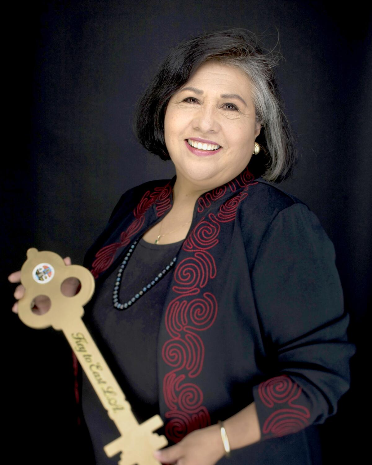In a waist-up frame, Gloria Molina poses for a portrait, smiling. She holds a prop that reads "Key to East L.A." 