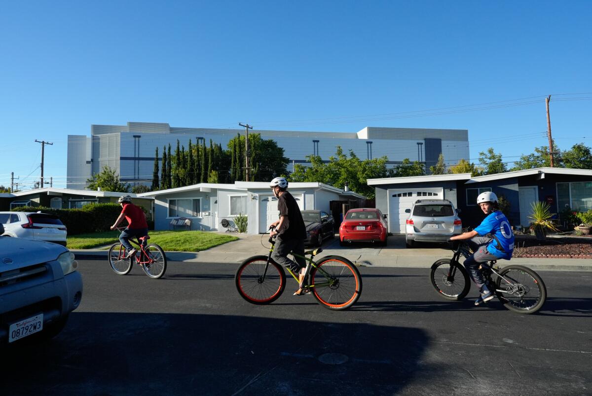 Boys ride bikes near homes and a large building in the background.