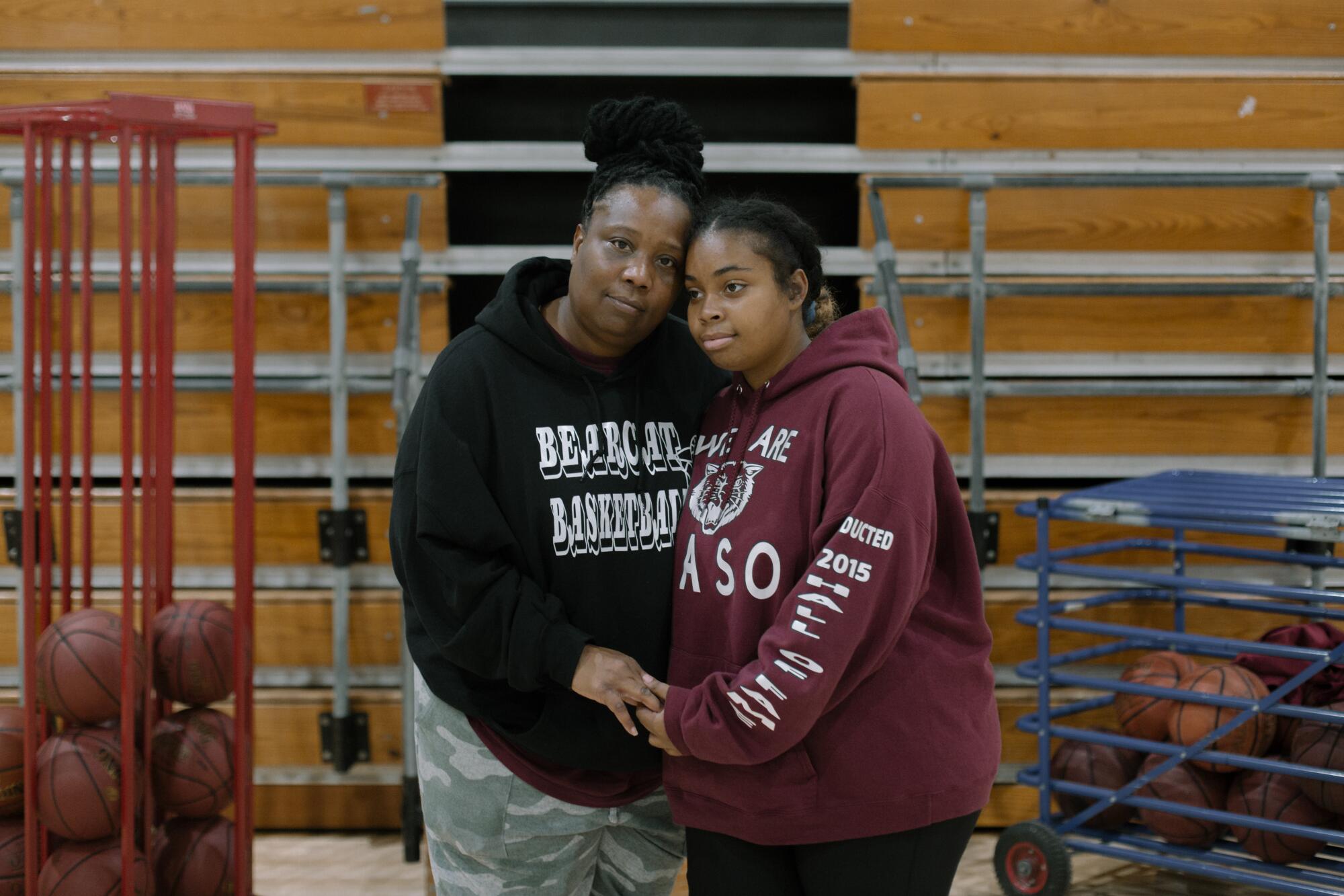 A woman and girl in Paso Robles High Bearcats sweatshirts leaning in to each other next to cages of basketballs in a gym.