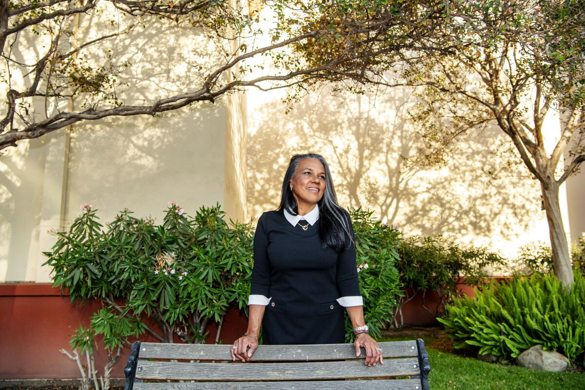 A woman stands behind a park bench in a lush outdoor setting.